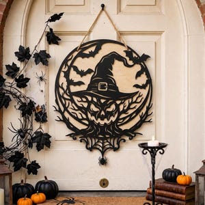 May include: A circular wooden wall hanging featuring a black laser-cut design of a witch hat, a jack-o'-lantern face, and flying bats. The piece is suspended by a tan rope and displays intricate branch and spiderweb patterns at the bottom.