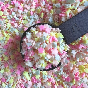 Könnte beinhalten: Nahaufnahme von bunten, blütenförmigen Streuseln in Rosa, Gelb, Weiß und Hellblau. Ein schwarzer Messlöffel hält einen Teil der Streusel, der Rest ist verstreut. Die Streusel sind wahrscheinlich zum Dekorieren von Backwaren gedacht.