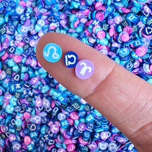 Può includere: Primo piano di tre piccoli ciondoli rotondi con segni zodiacali, tenuti su un dito. I ciondoli sono azzurri, blu scuro e lavanda, ognuno con un simbolo zodiacale bianco. Lo sfondo è un mix di coriandoli blu, viola e rosa a tema zodiacale.