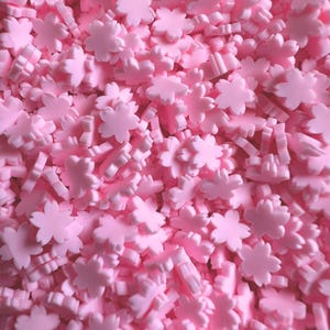 Puede incluir: Primer plano de un montón de pequeños adornos en forma de flor de color rosa claro. Las flores están dispersas y superpuestas, creando una superficie texturizada. Estos elementos decorativos son ideales para manualidades y proyectos de bricolaje.