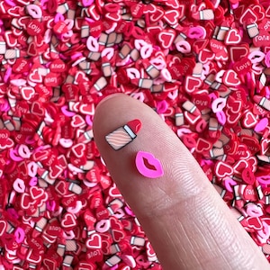 Peut inclure: Gros plan d'un doigt tenant deux breloques en pâte polymère : un rouge à lèvres et une forme de lèvre rose. L'arrière-plan est rempli de breloques en forme de cœur rouges et roses, certaines avec le mot "LOVE".