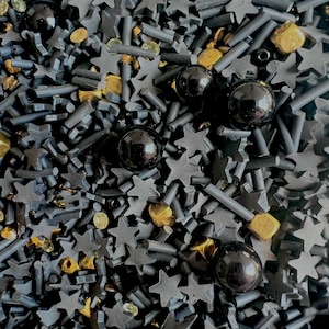Pode incluir: Close-up de uma mistura de granulados pretos em forma de estrela e bastão, intercalados com contas douradas e transparentes e granulados esféricos pretos. Os granulados estão espalhados, criando um efeito decorativo texturizado.