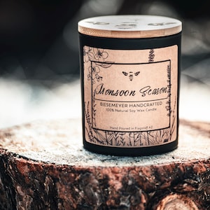 May include: A black candle jar with a wooden lid and a brown paper label that reads "Monsoon Season BIESEMEYER HANDCRAFTED 100% Natural Soy Wax Candle Hand Poured in Flagstaff AZ".