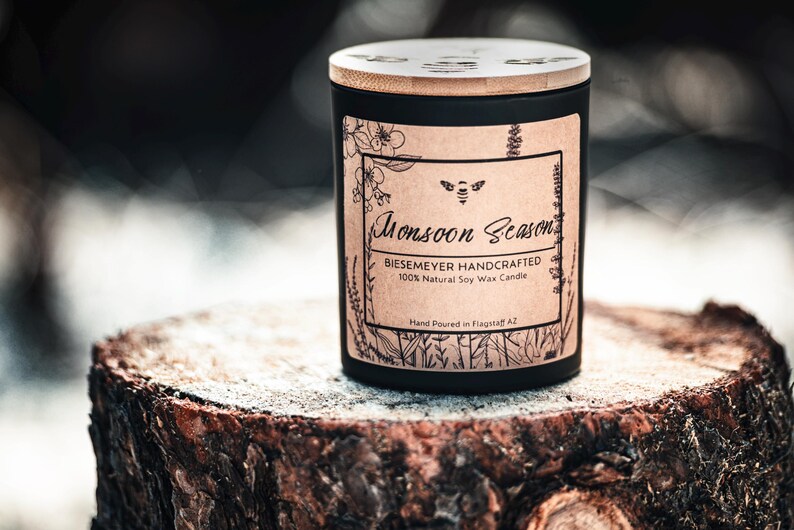 May include: A black candle jar with a wooden lid and a brown paper label that reads "Monsoon Season BIESEMEYER HANDCRAFTED 100% Natural Soy Wax Candle Hand Poured in Flagstaff AZ".