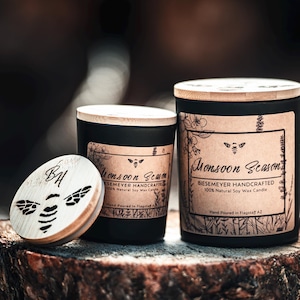May include: Two black soy wax candles with wooden lids. The larger candle has a label that reads "Monsoon Season BIESEMEYER HANDCRAFTED 100% Natural Soy Wax Candle Hand Poured in Flagstaff, AZ". The smaller candle has a wooden lid with the initials "BA" and a bee design.