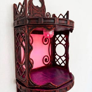 May include: A dark brown, gothic-style wall shelf with intricate cut-out designs and a pink interior. The shelf has a small, round shelf and a decorative top. The text "CRIMSON RELIQUARY" is printed at the bottom.