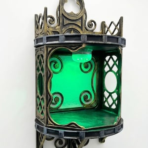 May include: A decorative corner shelf with an ornate, gothic design. The shelf is primarily bronze and black, with a green interior and a green light. The shelf has a curved front and is mounted on a white wall.