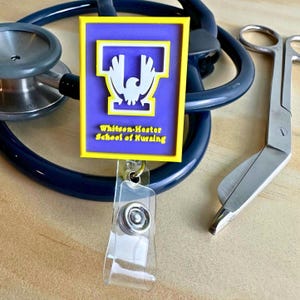 May include: A badge reel with a purple and yellow rectangular design featuring the text "Whitson-Hester School of Nursing". The badge reel is attached to a stethoscope and a pair of silver medical scissors.