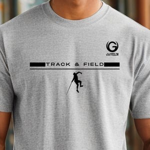 May include: A light gray t-shirt featuring the text "TRACK & FIELD" in black, with a silhouette of a javelin thrower. The word "JAVELIN" is above the text, with a stylized "G" logo. The shirt is a crew neck.