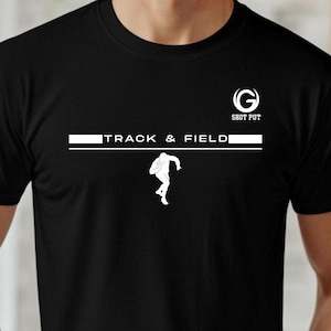 May include: Black t-shirt featuring a white graphic. The design includes the text "TRACK & FIELD" above a silhouette of a person throwing a shot put. The words "SHOT PUT" and a stylized "G" are at the top.