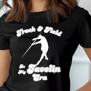 May include: A black short-sleeved t-shirt featuring a white graphic of an athlete throwing a javelin. The text "Track & Field" appears above the athlete, while the text "In My Javelin Era" is printed below in a bold, retro-style white font.