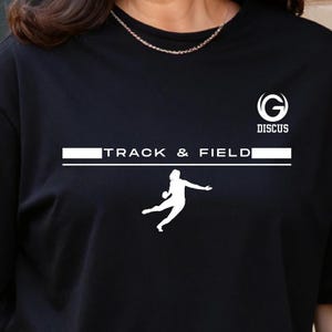 May include: Black t-shirt with white text and graphics. The shirt features the words "TRACK & FIELD" in a horizontal line, with a silhouette of a discus thrower below. The upper right corner has the word "DISCUS" and a stylized "G" logo.