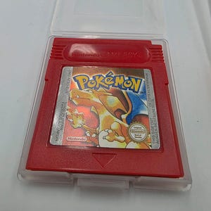 May include: A red Pokemon Game Boy cartridge in a clear plastic case. The cartridge features the Pokemon logo and a graphic of Charizard. The text "Nintendo Game Boy" is visible at the top.