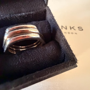 May include: A silver-toned ring with a multi-band design featuring three distinct, stacked horizontal ridges. The ring is nestled inside a dark brown, velvet-lined jewelry box. The partial text NKS is visible on a light-colored surface nearby.