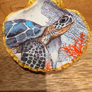 May include: A decorated scallop shell with a painted sea turtle design. The turtle has a blue and black shell with a detailed head. The shell is edged with gold foil and includes coral accents.
