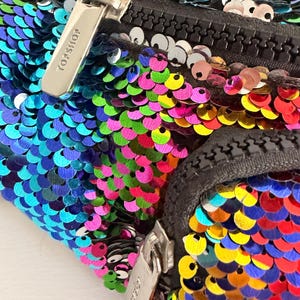Può includere: Un marsupio Topshop ricoperto di paillettes iridescenti multicolori nelle tonalità del blu, verde, rosa, giallo e arancione. La borsa presenta due scomparti neri con cerniera e tiretti in metallo argentato, ciascuno inciso con il logo Topshop.
