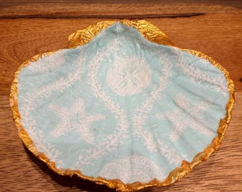 Puede incluir: Un plato decorativo en forma de concha con un interior azul claro con un patrón de estrellas de mar y algas blancas. El borde está adornado con pintura dorada, creando un tema costero.