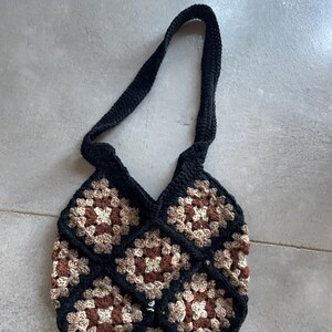 May include: A handmade crochet tote bag with a black strap and body. The body of the bag is made of brown, tan, and black granny squares. The bag is designed to be carried over the shoulder.