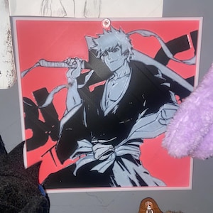 May include: A framed anime art print depicting a male character with spiky, light blue hair and a black outfit, holding a sword. The background is a solid red colour. The art style is a bold, graphic illustration.