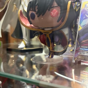 May include: A stylized vinyl figure of a character with black hair, wearing a black and gold high-collared cape with a red interior. The figure features one purple eye and one red eye with a bird-like symbol, standing on a clear circular base.