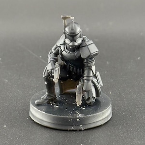 May include: A black and gray plastic action figure of a Star Wars clone trooper, wearing a helmet and holding a blaster. The figure is posed in a crouching position and is standing on a black plastic base.