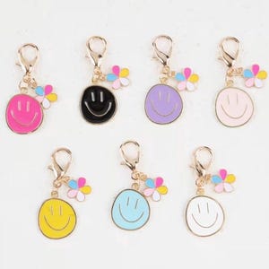 May include: A collection of seven smiley face charms in various colors, including pink, black, purple, light pink, yellow, blue, and white. Each charm has a gold-colored clasp and a small flower accent with pastel petals.