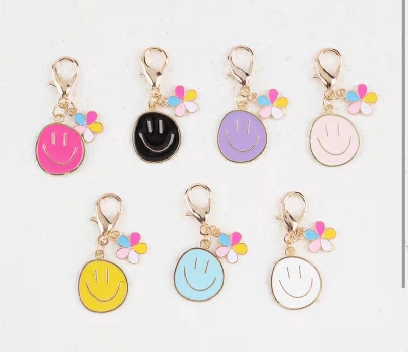 May include: A collection of seven smiley face charms in various colors, including pink, black, purple, light pink, yellow, blue, and white. Each charm has a gold-colored clasp and a small flower accent with pastel petals.