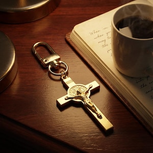 May include: A gold crucifix keyring featuring a detailed depiction of Jesus Christ. The cross is attached to a gold clasp. The keyring rests on a dark wooden surface next to a book and a cup of coffee.