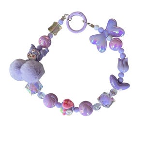 Pu&ograve; includere: Un braccialetto stravagante nei toni del lavanda, lilla e bianco iridescente. Il braccialetto presenta una variet&agrave; di perline, tra cui elementi rotondi, quadrati e a forma di fiore. Un soffice pompon lavanda e un ciondolo a forma di personaggio dei cartoni animati aggiungono un tocco giocoso.