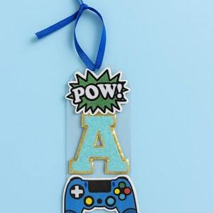 May include: A gaming-themed bookmark with a blue ribbon. The bookmark features the word "POW!" in a comic book style, a teal letter "A" with gold trim, and a blue video game controller. The background is light blue.