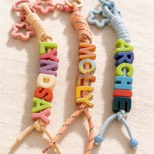 Puede incluir: Tres llaveros personalizados con nombres escritos con cuentas de letras coloridas. Los nombres son "LYNDSAY", "MOLLY" y "ARCHIE". Cada llavero tiene una combinación de colores diferente y dijes en forma de estrella o redondos.