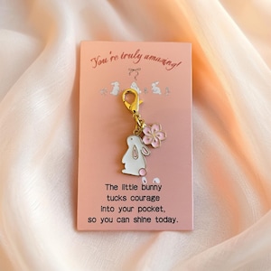 Puede incluir: Una tarjeta rosa con el texto "You're truly amazing!" y un dije con un conejito blanco y una flor rosa. El dije está sujeto a la tarjeta con un cierre dorado. La tarjeta también tiene el texto "The little bunny tucks courage into your pocket, so you can shine today."