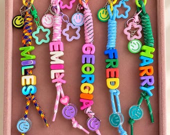 May include: Five colorful keychains featuring braided cords in various patterns and colors. Each keychain includes plastic letter beads spelling the names MILES, EMILY, GEORGIA, FRED, and HARRY, accented with small smiley face charms and star-shaped plastic links.