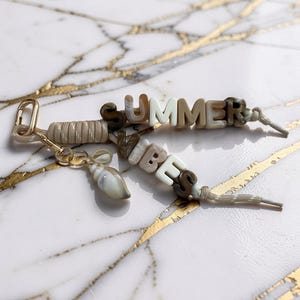 以下が含まれることがあります： 「SUMMER VIBES」の文字がニュートラルカラーの文字ビーズで綴られたビーズキーホルダー。小さな貝殻チャームとゴールドカラーの留め金が付いており、鍵やバッグに取り付けるのに最適です。