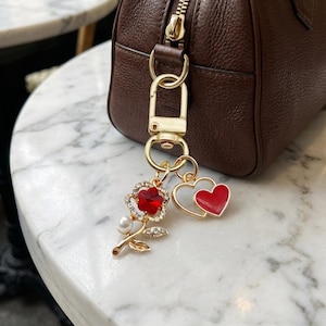 May include: A gold-toned keychain charm featuring a red rose with rhinestone accents and a pearl, plus a red heart charm. The charms are attached to a gold clasp, hanging near a brown leather handbag.