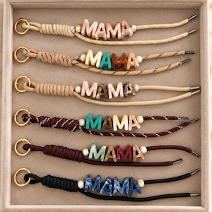 May include: Six bracelets featuring the word MAMA spelled out in marbled acrylic letters. Each bracelet has a gold-toned metal ring, a cord section with a wrapped design, and two small white beads flanking the lettered name on a cord.