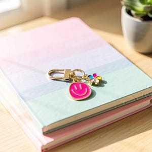 May include: A stack of pastel-colored notebooks with a gold keychain featuring a pink smiley face charm and a flower charm. The notebooks are pink, blue, and green, and rest on a wooden surface.