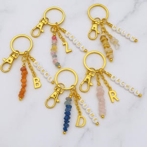 Peut inclure: Porte-clés dorés avec fermoirs mousquetons, personnalisés avec des noms épelés en perles blanches. Chaque porte-clés comprend un charme de pierres précieuses colorées et une lettre initiale dorée. Les porte-clés sont présentés sur une surface en marbre blanc.