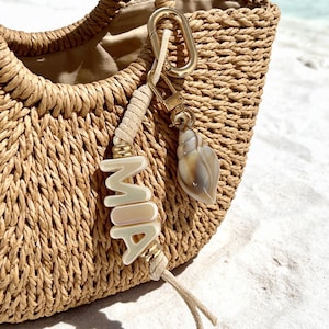 Könnte beinhalten: Eine geflochtene Strohtasche mit einem dekorativen Schlüsselanhänger. Dieser besteht aus einer beigen Kordel mit goldfarbenen Perlen, weißen Buchstabenperlen, die MIA bilden, und einem natürlichen braun-weißen Muschelanhänger an einem goldenen Verschluss.