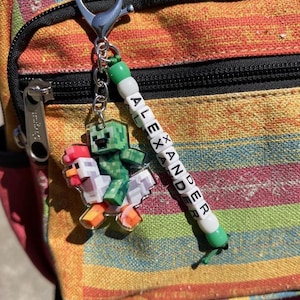 May include: A keychain featuring a green pixelated Creeper character riding on two pixelated chickens. The keychain includes a beaded name tag that spells "ALEXANDER" in white letter beads. The keychain is attached to a silver clasp.