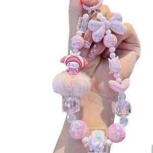Pu&ograve; includere: Un portachiavi rosa con una variet&agrave; di elementi decorativi. Presenta un personaggio dei cartoni animati, un soffice pompon, perline iridescenti, un fiocco e accenti a forma di cuore. Il portachiavi &egrave; tenuto da una persona.