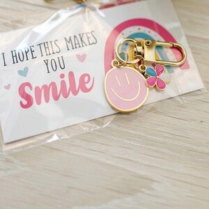 May include: A gold-colored keychain with a pink smiley face charm and a flower charm with pink, blue, and white petals. The keychain is attached to a card that reads "I HOPE THIS MAKES YOU SMILE" with a rainbow graphic.