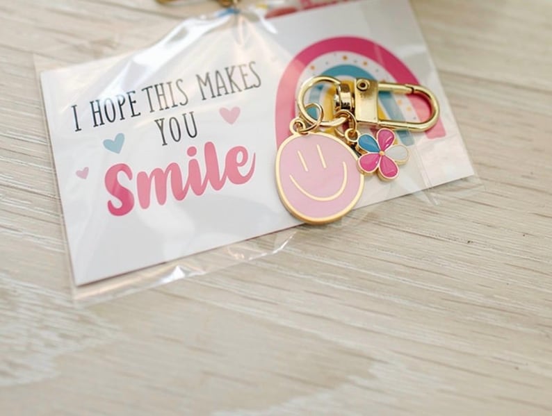 May include: A gold-colored keychain with a pink smiley face charm and a flower charm with pink, blue, and white petals. The keychain is attached to a card that reads "I HOPE THIS MAKES YOU SMILE" with a rainbow graphic.