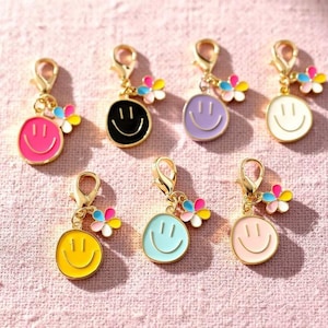 May include: A collection of colorful smiley face charms with gold-colored clasps. Each charm features a different color smiley face, including pink, black, purple, white, yellow, and light blue, with a small flower accent. The charms are likely for jewelry or accessories.
