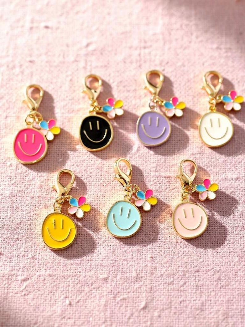 May include: A collection of colorful smiley face charms with gold-colored clasps. Each charm features a different color smiley face, including pink, black, purple, white, yellow, and light blue, with a small flower accent. The charms are likely for jewelry or accessories.