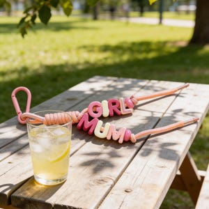 May include: A wooden picnic table holds a glass of lemonade with ice and a lemon slice. Two pink and peach rope keychains spell out "GIRL MUM" in colorful letters. A pink carabiner clip is attached to one keychain.