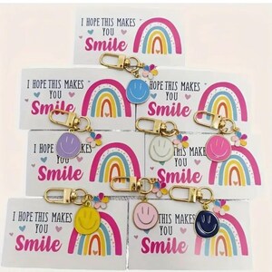 May include: Keychains with colorful smiley faces in blue, purple, white, pink, yellow, and navy blue. Each keychain has a gold clasp and a small flower charm. The keychains are displayed with cards that read "I HOPE THIS MAKES YOU Smile".