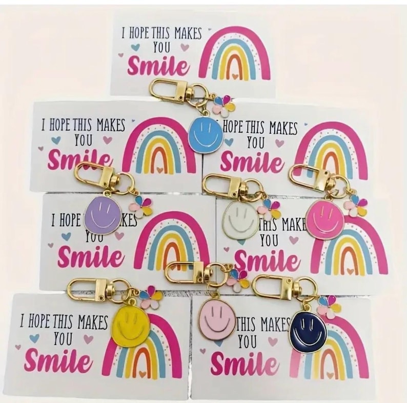 May include: Keychains with colorful smiley faces in blue, purple, white, pink, yellow, and navy blue. Each keychain has a gold clasp and a small flower charm. The keychains are displayed with cards that read "I HOPE THIS MAKES YOU Smile".