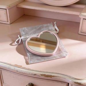 Peut inclure: Un miroir de poche rose en forme de cœur est posé sur un sac en organza argenté. Le miroir est sur une coiffeuse rose avec un miroir ovale assorti. La coiffeuse a des tiroirs et des détails ornés.