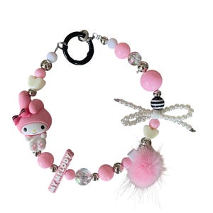 Pu&ograve; includere: Un braccialetto di perline rosa con un ciondolo My Melody, perline a forma di cuore e un pompon rosa. Il braccialetto include un ciondolo a forma di libellula e una barra con la scritta "MY MELODY". Il braccialetto ha una chiusura nera.